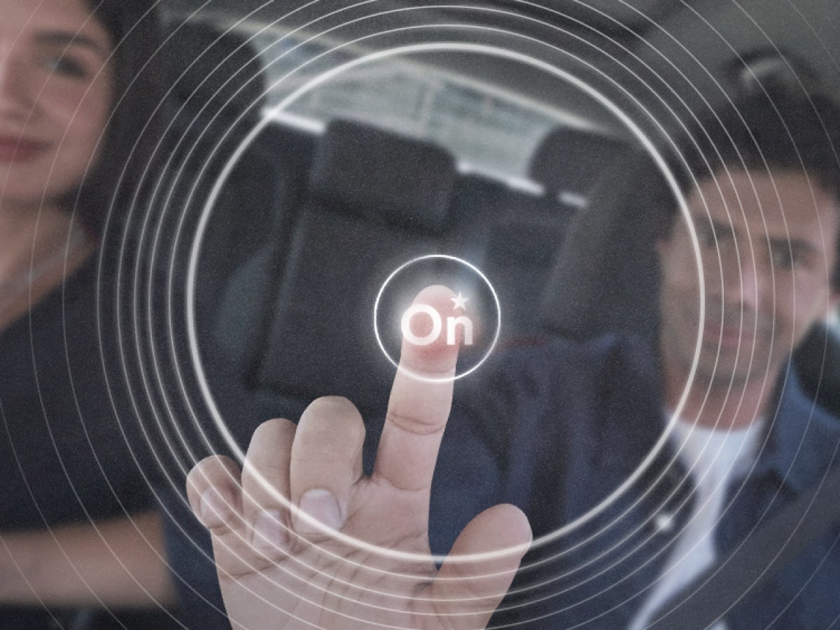 Primer plano de una mano presionando un botón digital /On/ dentro de un vehículo, con efecto visual de ondas que representan la activación de tecnología OnStar. Detrás aparecen un hombre y una mujer sentados en el vehículo.