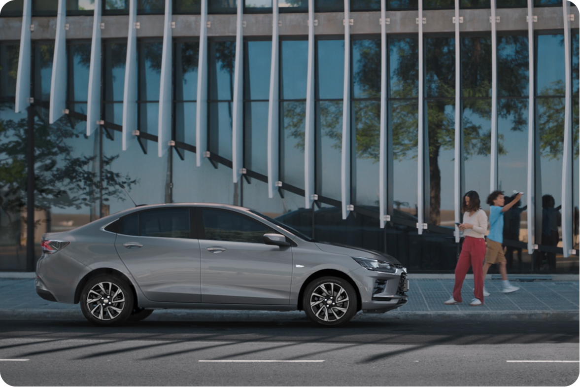 Chevrolet Onix Plus na cor Prata Shark estacionado em frente a fachada moderna, com duas pessoas caminhando ao lado.