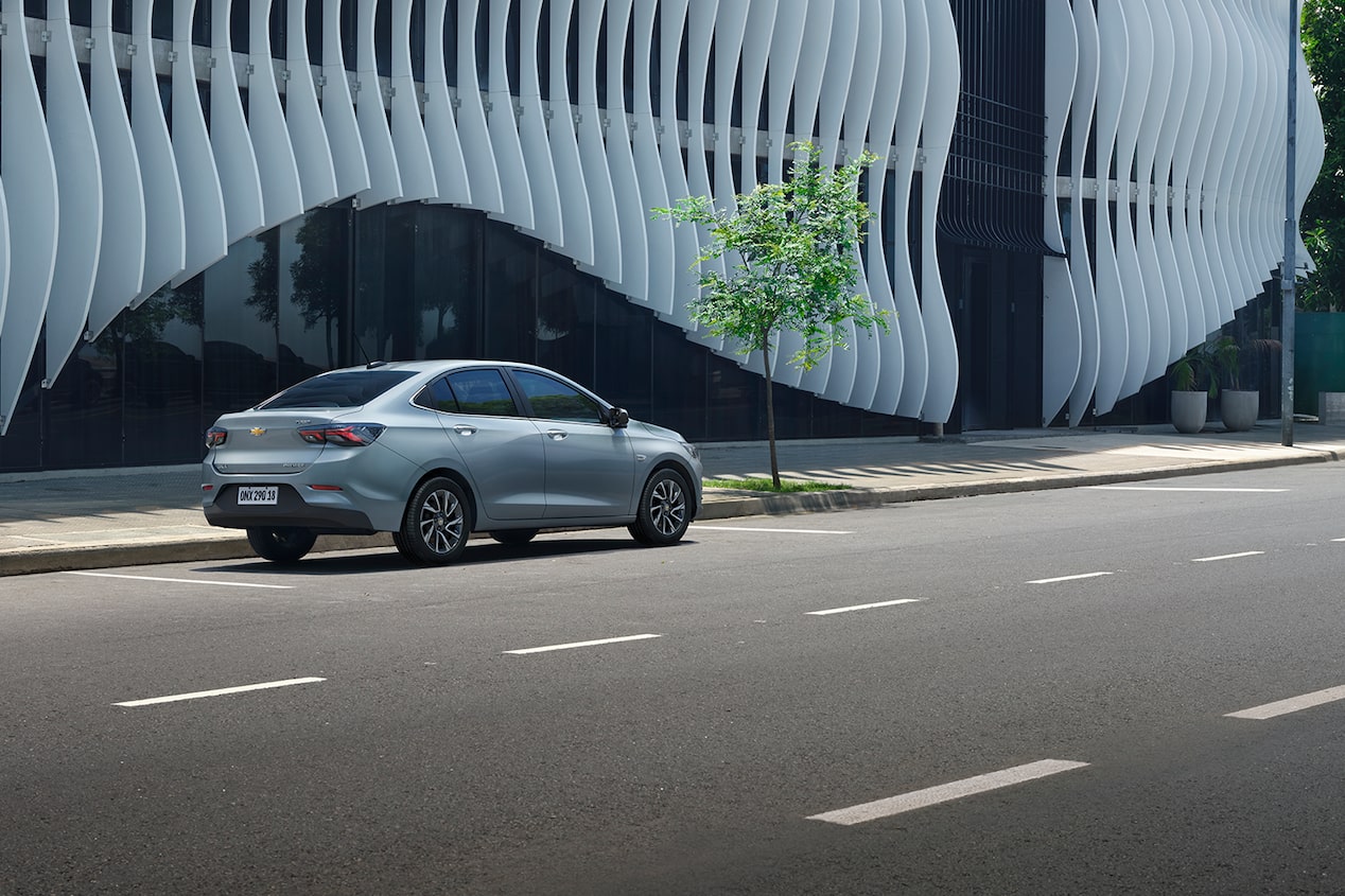 Chevrolet Onix Plus na cor Prata Shark circulando em avenida com fachada arquitetônica de linhas onduladas ao fundo.