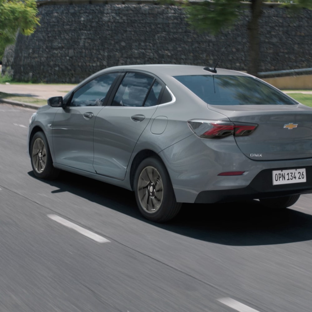 Vista traseira do Chevrolet Onix Plus na cor Prata Shark, em movimento com muro de pedra ao fundo.
