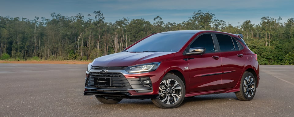 Masthead da página de acessórios originais Chevrolet, versão desktop, para o carro hatchback Onix, que aparece em foto ambientada, estacionado em frente a um paredão de árvores verdes.