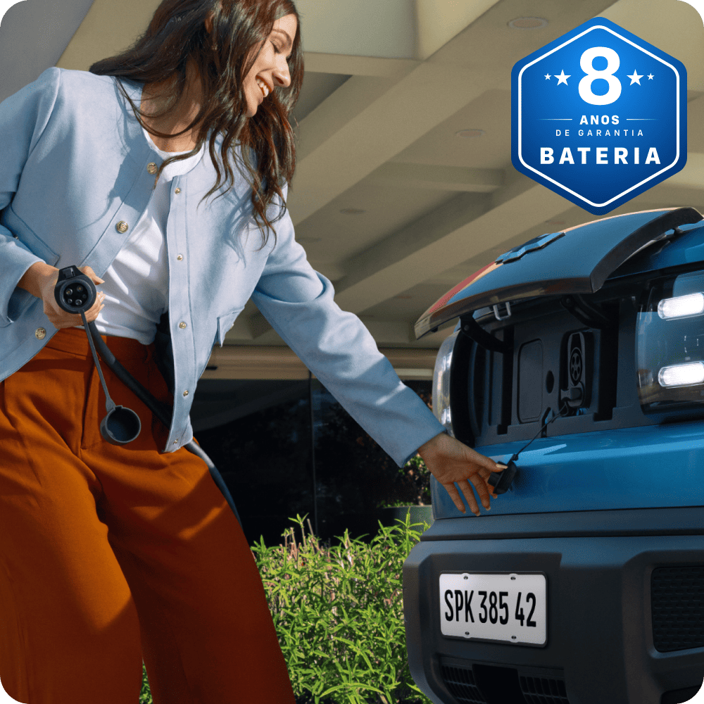 Mulher sorridente conecta o carregador ao Chevrolet Spark em ambiente residencial. Selo azul indica 8 anos de garantia da bateria.