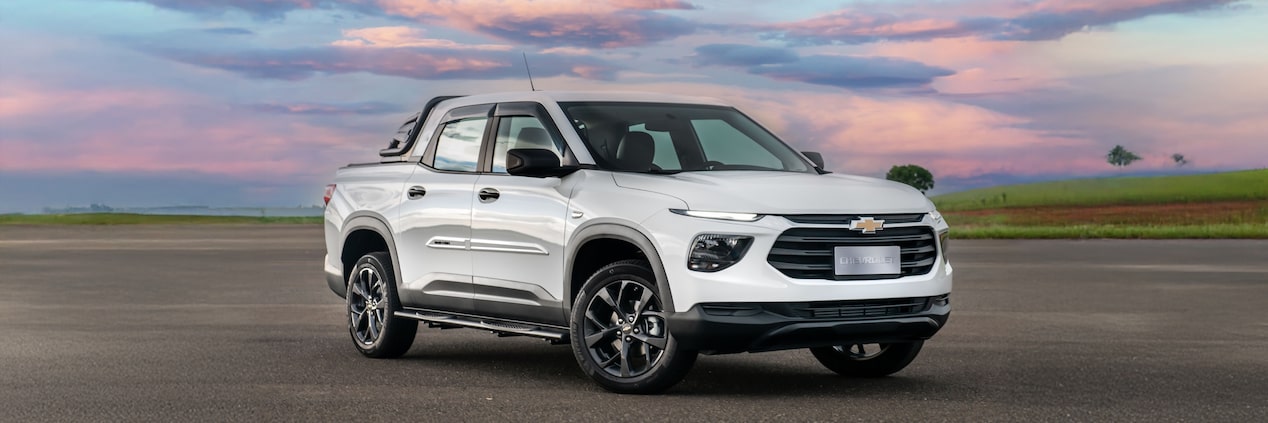 Chevrolet Montana branca em imagem ambientada, estacionada a céu aberto, em posição 3/4 de frente.