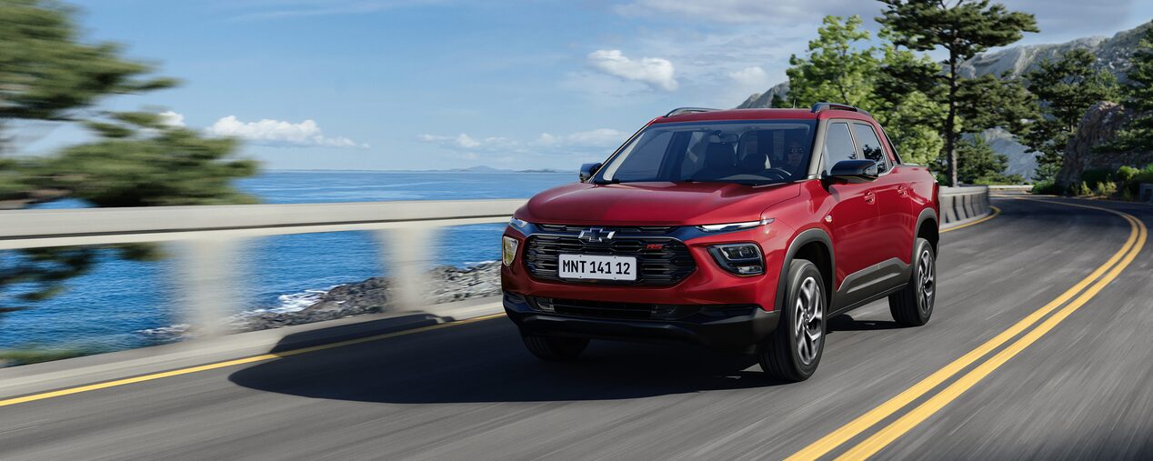 Chevrolet Montana versão RS na cor Vermelho Scarlet em rua asfaltada à beira-mar, com guarda-corpo branco, árvores e céu azul