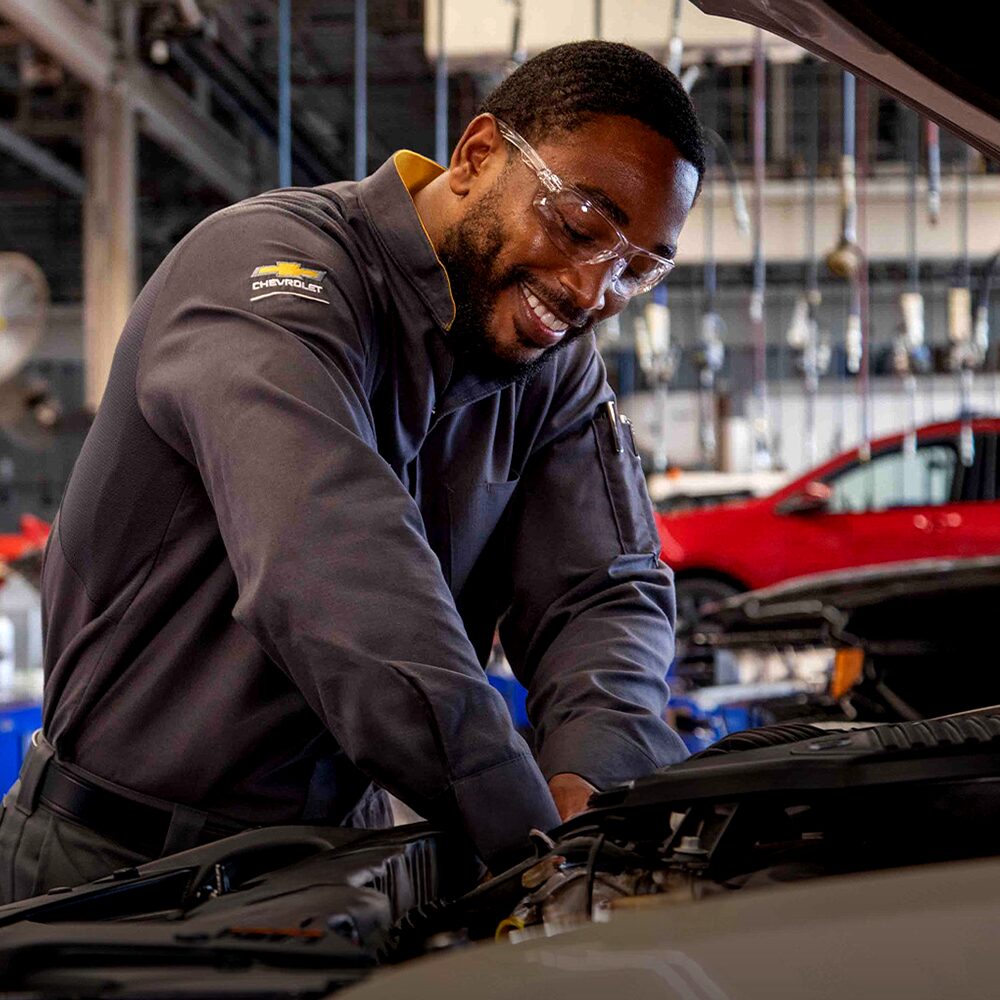 Mecânico Chevrolet sorridente, usando uniforme preto com logotipo da marca e óculos de proteção, realizando manutenção no motor de um veículo dentro da oficina.
