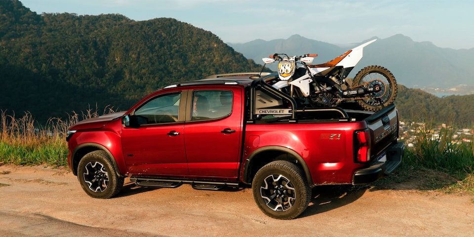 Chevrolet S10 Z71 2026 vermelha usada pelo  o influenciador Lucas Xaparral estacionada em local de terra batida à beira-mar. A caçamba está carregada com uma prancha de surfe e uma motocicleta. 