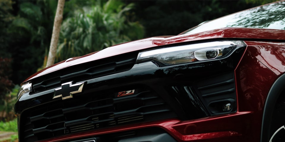 Close do farol e grade frontal da Chevrolet S10 Z71 2026 vermelha, com destaque para o emblema preto da Chevrolet e o logotipo Z71. O fundo mostra vegetação tropical, reforçando o clima de aventura.
