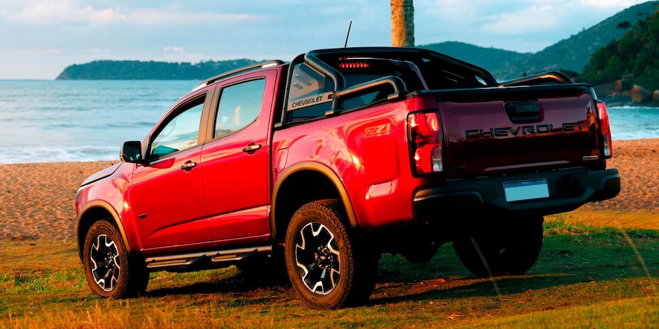 Chevrolet S10 Z71 2026 vermelha estacionada de costas na areia da praia, com mar e morros ao fundo. A imagem destaca a traseira do veículo, incluindo o nome Chevrolet em alto relevo e o santa antônio esportivo.
