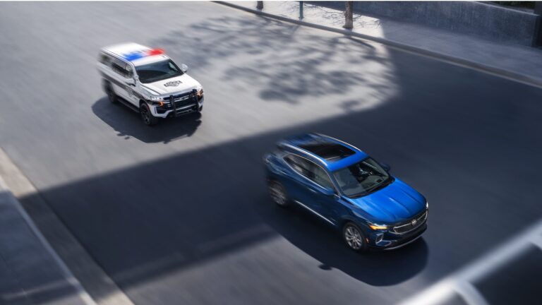 SUV Chevrolet azul sendo acompanhado por uma viatura policial em movimento em avenida iluminada pelo sol.