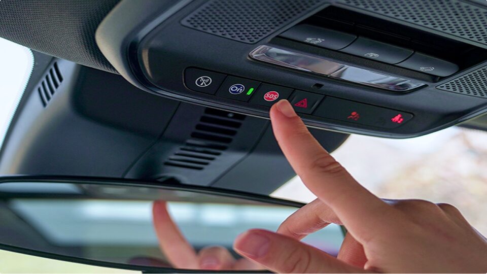 Mão de uma pessoa prestes a pressionar o botão vermelho de emergência do sistema OnStar, localizado no console superior próximo ao retrovisor interno do carro.