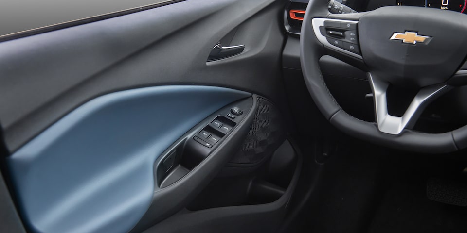 Interior del Chevrolet Onix Sedán visto desde el puesto del conductor, mostrando el volante con el emblema de Chevrolet y el panel de la puerta con detalles en azul y gris en los controles eléctricos de las ventanillas.